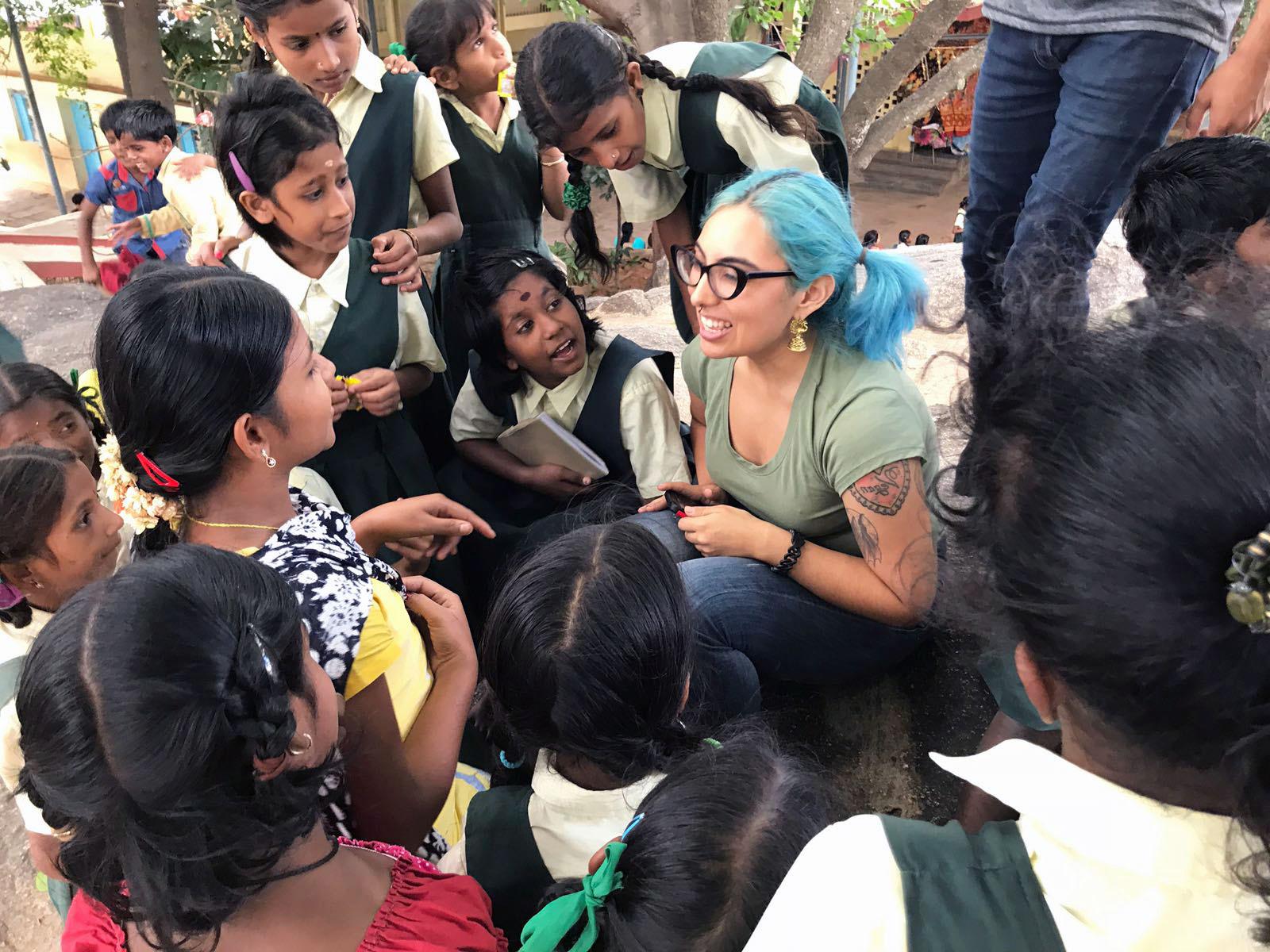 Smiling Cal State LA student sitting on the ground surrounded by listening young school students in India.